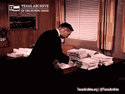 A man in a suit standing over a desk covered in tall stacks of documents, working through them one at a time